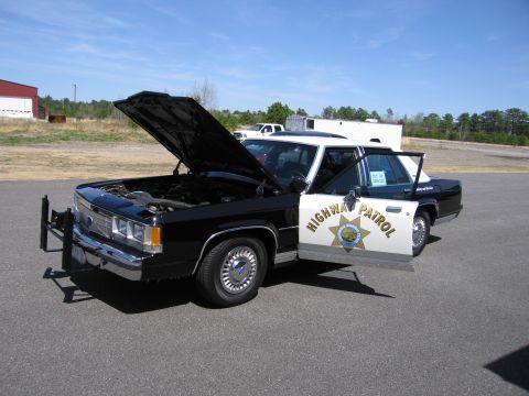1991 Ford LTD Crown Victoria - A black and white Ford Crown Victoria police car is parked with its hood and driver's door open under a clear blue sky.