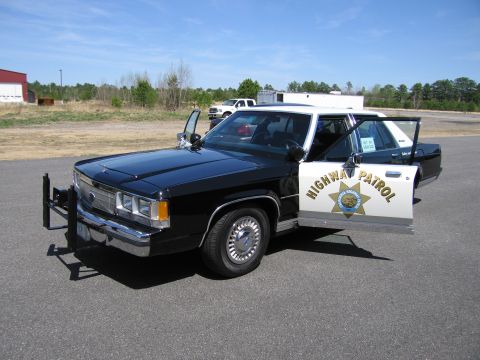 1991 Ford LTD Crown Victoria - A black and white highway patrol car is parked on asphalt with its doors open under a clear blue sky.