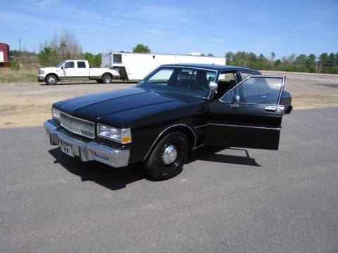 1991 Ford LTD Crown Victoria - This black sedan, likely from the 1980s, is parked outdoors with its driver's door ajar. A white pickup truck and trailer are visible in th…
