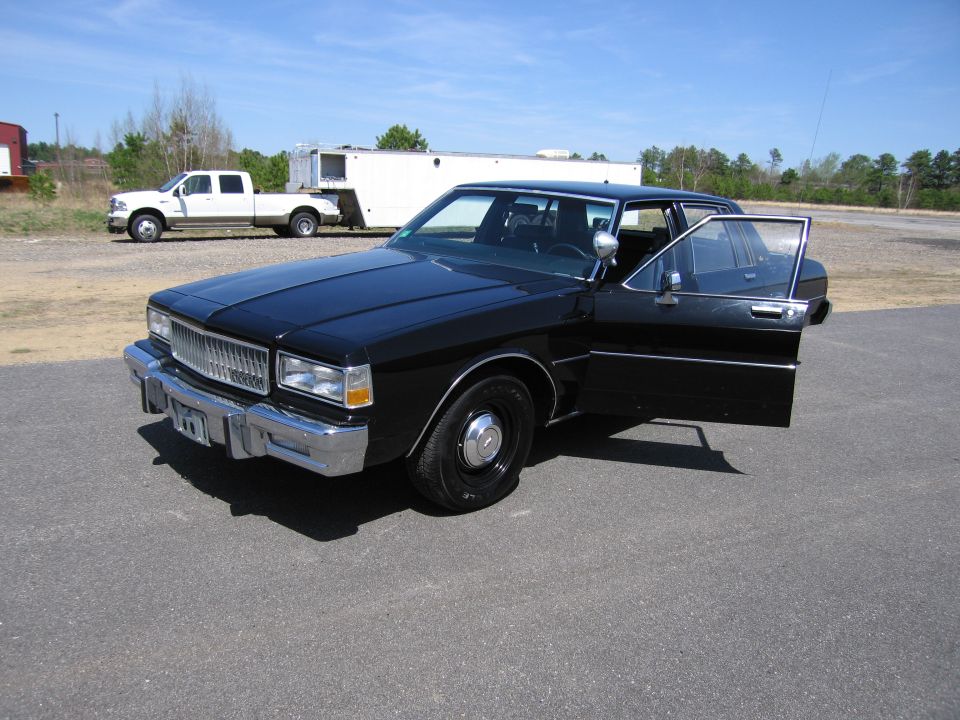Black sedan, driver's door open, parked on asphalt with a white pickup truck and trailer in the background