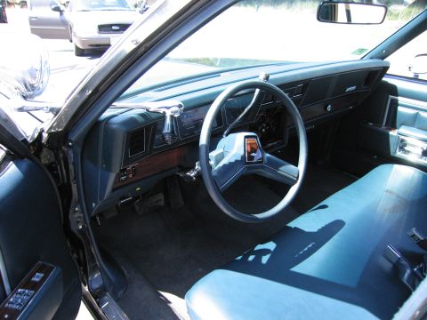 1991 Ford LTD Crown Victoria - Interior view of a car, showing the dashboard, steering wheel, and blue bench seat. Another vehicle is visible through the window.