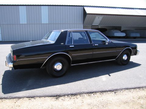 1991 Ford LTD Crown Victoria - A black sedan is parked on asphalt in front of a metal building with covered vehicles in the background.