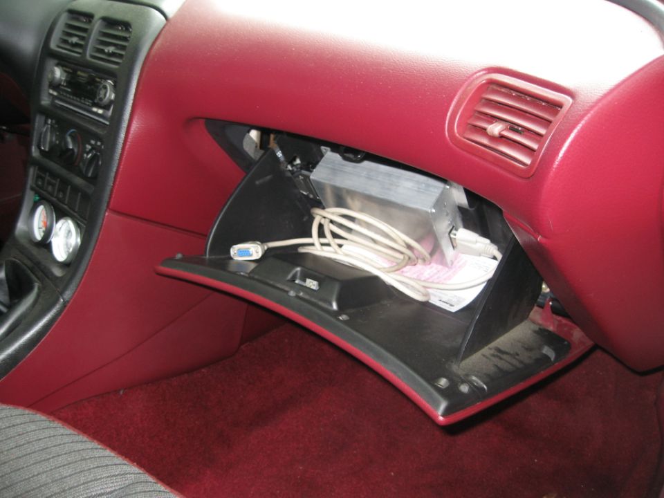 Car interior showing an open glove box with wires and a metal box inside. Red dashboard and carpet visible