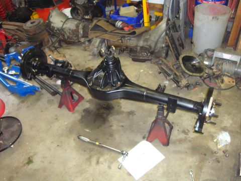 1989 Ford Bronco - A black rear axle assembly is supported by red jack stands in a workshop environment with various automotive parts scattered around.