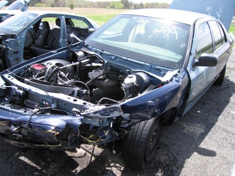 2001 Ford Crown Victoria - A damaged police car sits in a junkyard, with its hood open and front end heavily impacted.