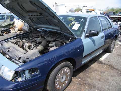 2001 Ford Crown Victoria - A two-tone blue police car, possibly a 2001 Ford Crown Victoria, sits with its hood open in a junkyard.