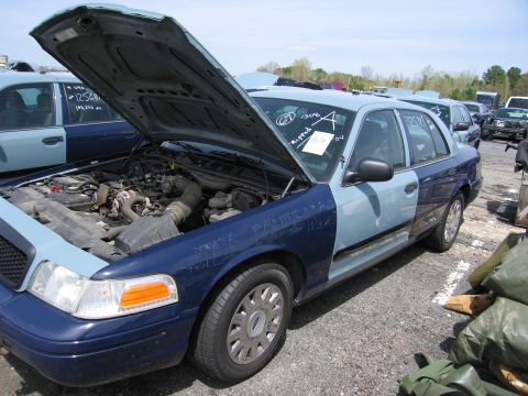 2001 Ford Crown Victoria - A two-tone blue 2001 Ford Crown Victoria sits in a junkyard with its hood open, revealing the engine compartment.