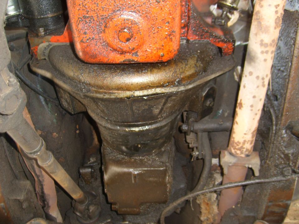 Underside view of an engine with an oil pan and a rusty exhaust pipe. Lots of grease and grime visible