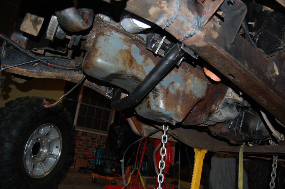 Underside view of a vehicle's engine and frame, with a large tire visible on the left. It appears to be undergoing maintenance or repair