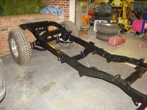 1948 Jeep Wrangler - A freshly painted black frame chassis sits on a garage floor with front axle and two off-road tires mounted, viewed from the front-left thr…