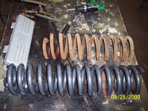 1979 Ford Bronco - Comparison of a rusty old coil spring with a new gray one, alongside tools and a metal component on a workbench.