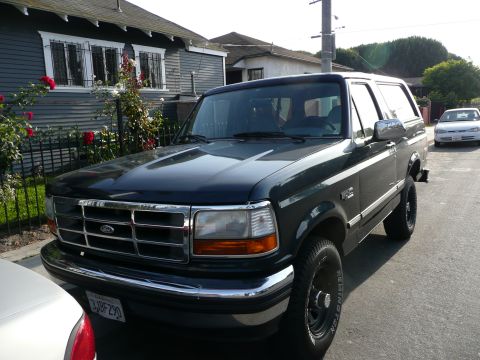 1991 Ford Bronco - Dark green Ford Bronco parked on a street with a house and fence in the background.