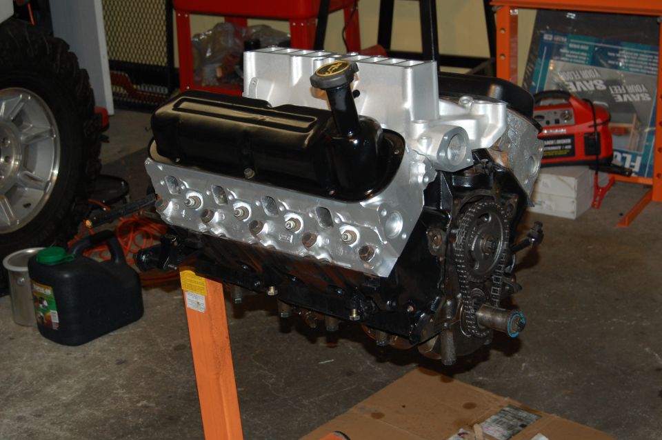 Engine on an orange stand, with a black valve cover and silver intake manifold. A tire and a green gas can are visible to the left