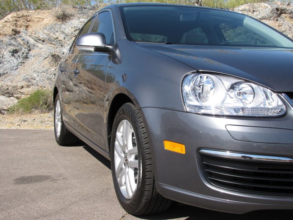 Passenger-side front corner of a gray VW Jetta with clear headlight and alloy wheel, desert rocks behind