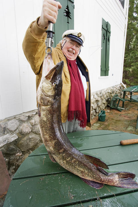 Man in a yellow jacket and red scarf holds a large fish by its mouth, with a white building in the background