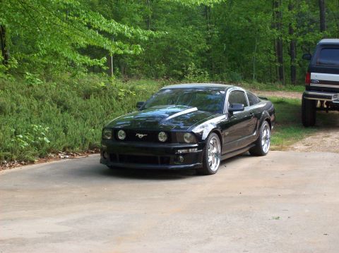 2007 Ford Mustang - A black Ford Mustang is parked on a driveway next to a dark SUV, with trees in the background.