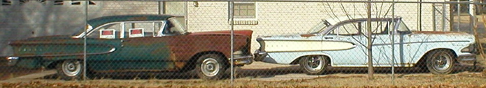 Two weathered 1950s Edsel sedans parked behind a chain-link fence, one green and one pale blue.