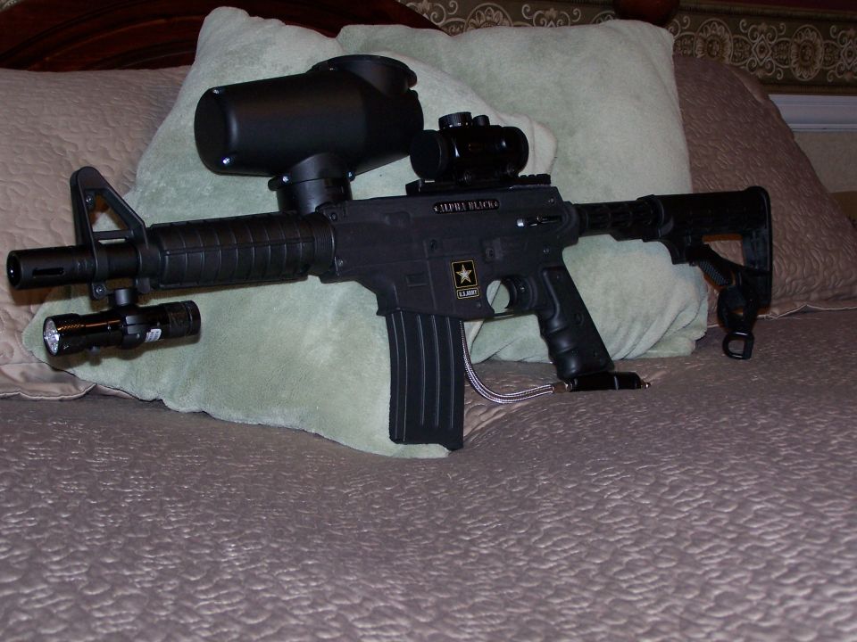Black AR-style marker with red dot sight and flashlight resting on a green pillow on a bed