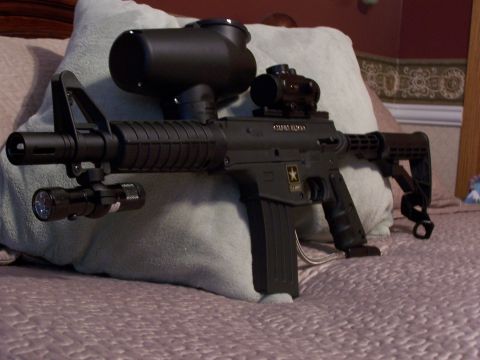 1986 Ford Bronco - A black paintball gun is shown with a tactical flashlight and red dot sight attached, resting on a bed.
