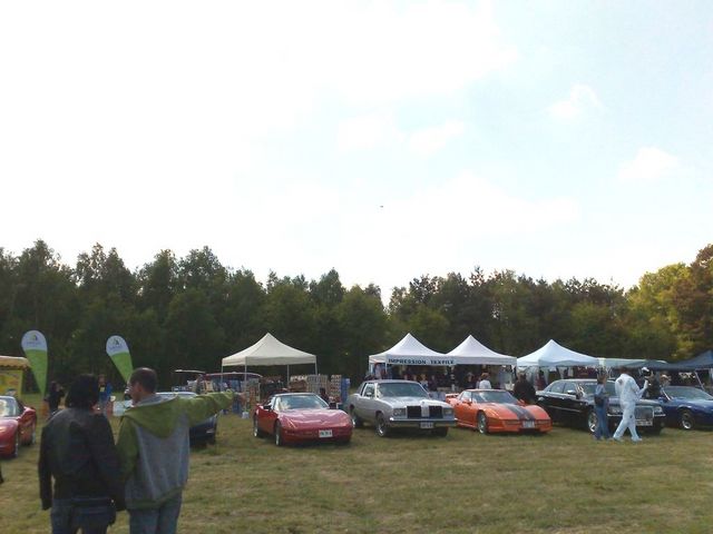 Outdoor car show with tents set up, several cars parked on grass, and people milling around