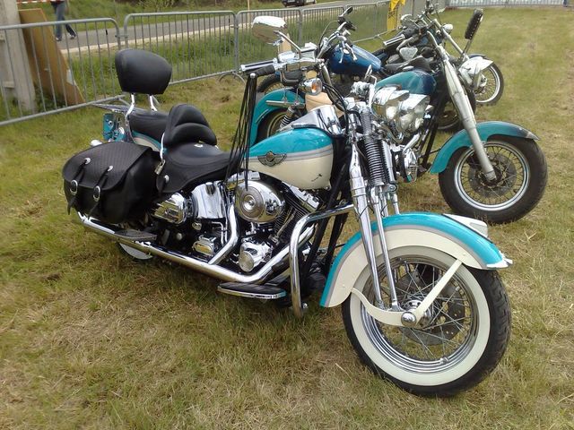 Two motorcycles parked on grass, one in foreground with black saddlebags and two-tone teal and cream body