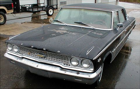 1983 Ford Bronco - A classic black Ford sedan sits on a wet lot, with a Ford Bronco and trailer visible behind it.