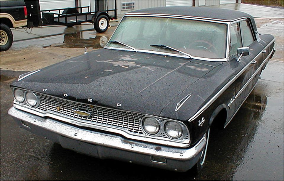 Black early-1960s Ford Galaxie sedan, front 3/4 view, wet paint on a lot.