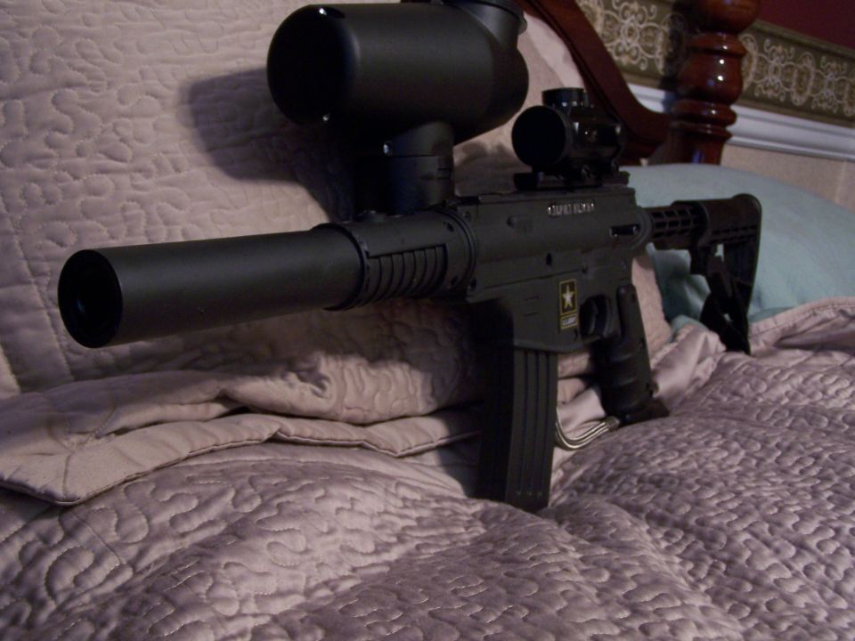 Black paintball gun with scope and suppressor, resting on a quilted bedspread