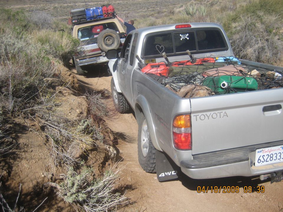 Silver Toyota Tacoma TRD pickup loaded with gear on a dirt trail behind a tan SUV