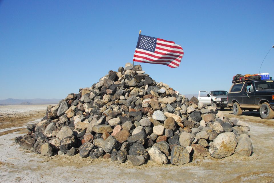 Rock cairn with American flag on a desert lakebed, loaded Ford Bronco parked at right