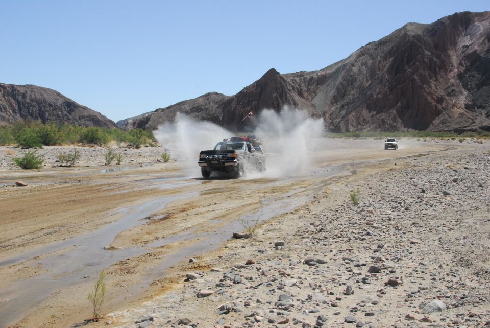 Dark Ford Bronco splashing through a desert water crossing with tall spray and mountains behind