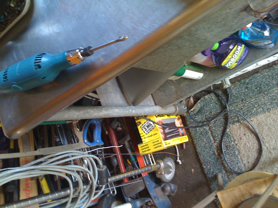Teal drill on metal bench above cluttered garage shelves with tools, wire, and cleaning products