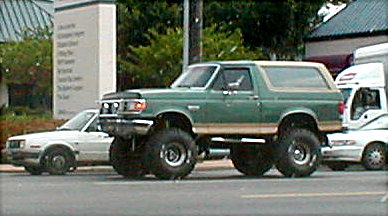 1983 Ford Bronco - A modified green Ford Bronco with large tires is parked on the street next to other vehicles.