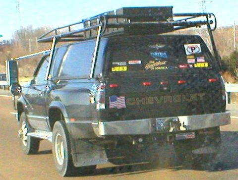 1983 Ford Bronco - Rear view of a black Chevrolet Suburban equipped with a roof rack and bed cap, adorned with various stickers.