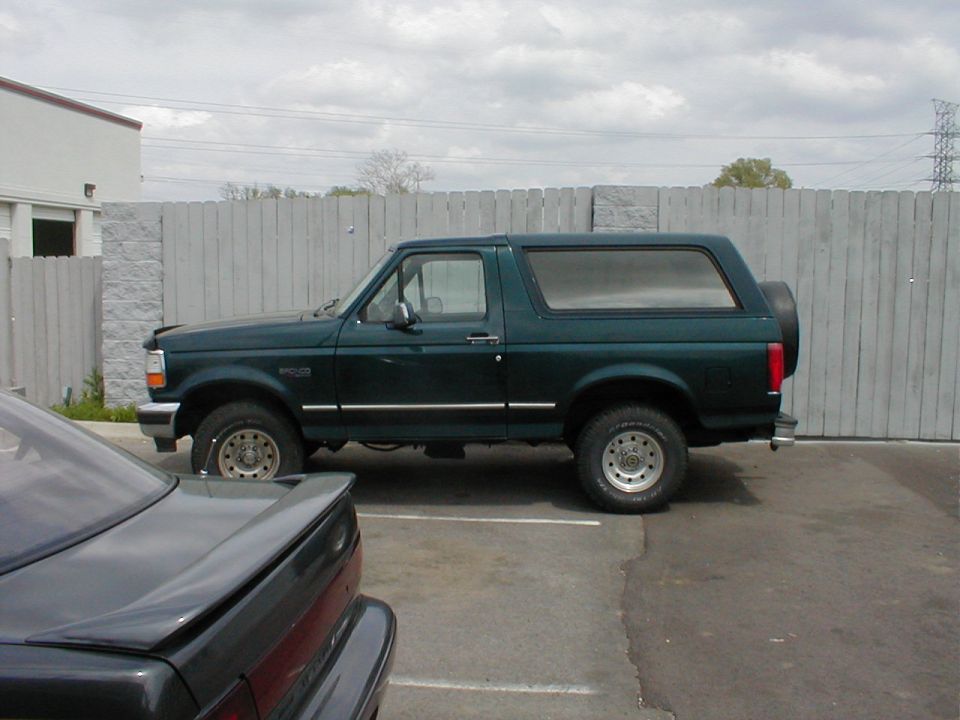 Dark green two-door Ford Bronco SUV, side view in a fenced parking lot.