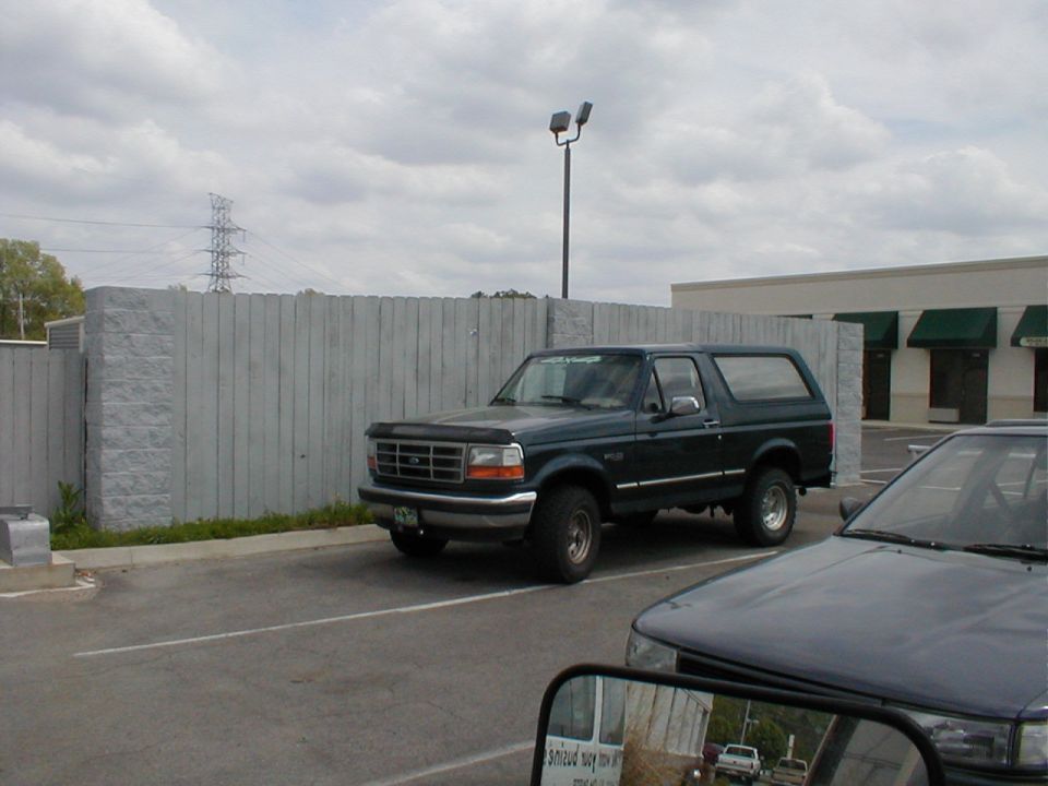 Dark green Ford Bronco SUV parked by a gray fence in a paved lot.