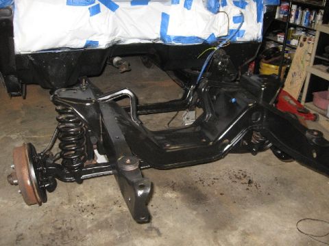 1966 Ford F-100 - Freshly painted gloss-black front frame and coil-spring suspension of a truck in a garage, with the bare rust-colored brake drum and hub ex…