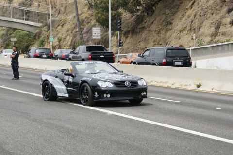 1993 Ford Taurus - A black Mercedes-Benz convertible with racing decals drives on a highway, with other vehicles and a police officer visible.