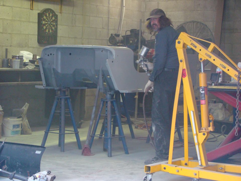 Man sprays primer on Jeep CJ-5 body shell on stands, yellow engine hoist nearby