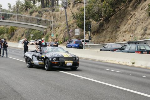 1993 Ford Taurus - A black convertible Mustang with gold racing stripes is escorted by police on a highway.