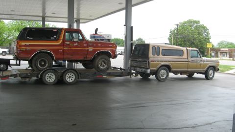 1979 Ford Bronco - A red 1979 Ford Bronco is transported on a trailer pulled by a brown pickup truck at a gas station.