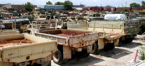 1983 Ford Bronco - A collection of aging military trucks are parked in rows at a salvage yard under a cloudy sky.