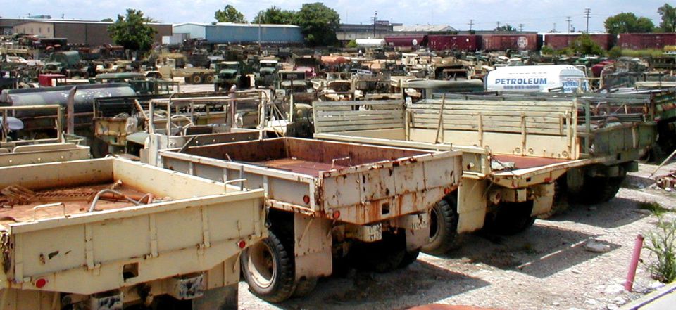 Rows of surplus military cargo trucks and trailers in a dirt storage lot
