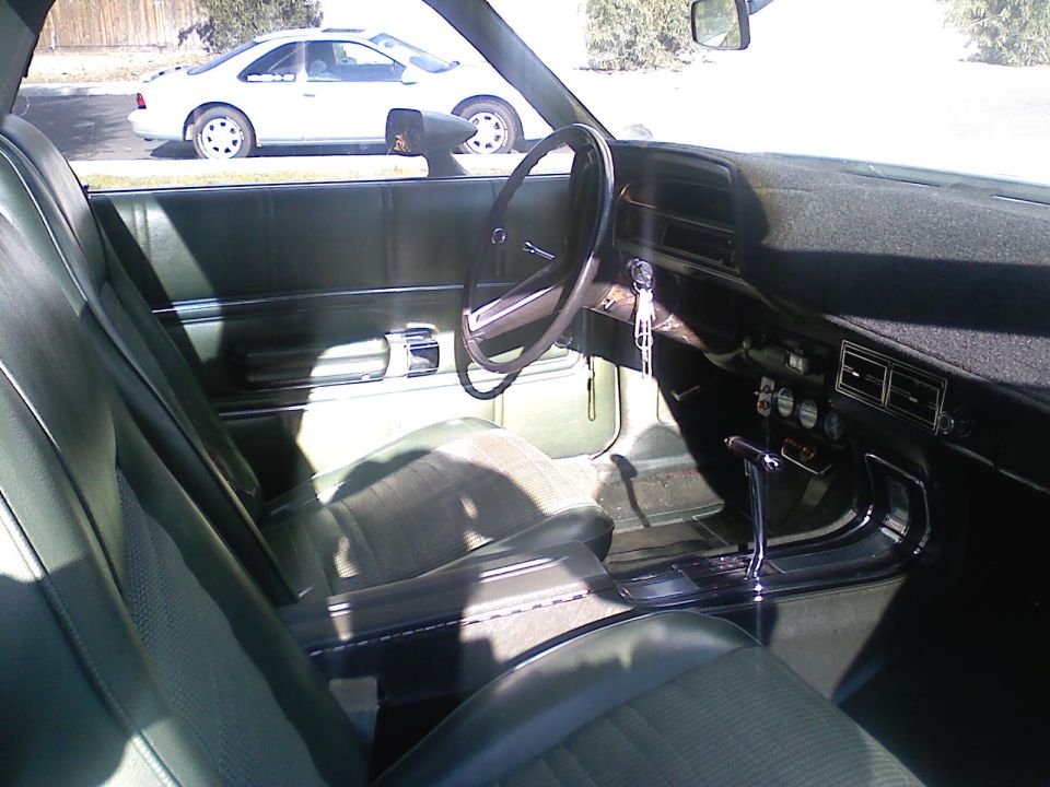 Green interior of a 1970s pickup truck or car, showing dashboard, steering wheel, seats, and gear shifter. Another white car is visible through the window