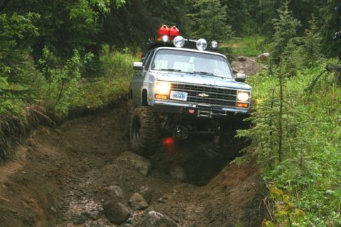 1990 Chevrolet Suburban - A lifted Chevrolet K5 Blazer navigates a steep, muddy off-road path surrounded by dense forest.