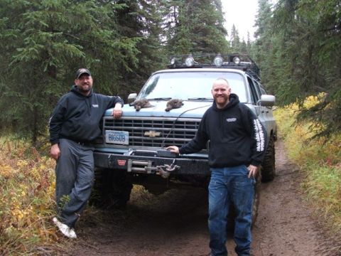 1990 Chevrolet Suburban - Two men pose with a modified Chevrolet K5 Blazer in a wooded area, suggesting an off-road adventure.