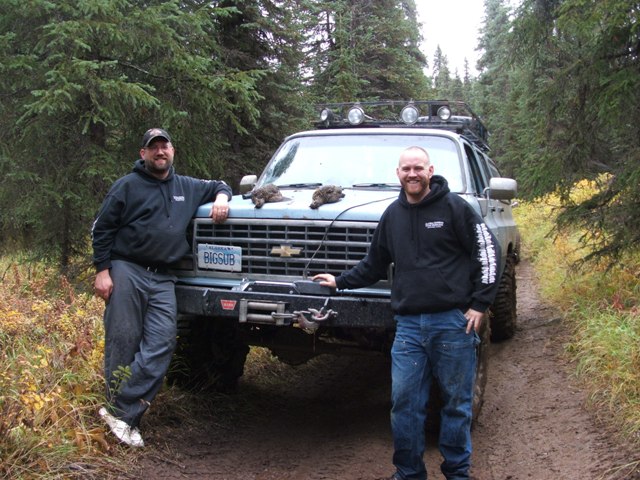 Two guys stand by a lifted K5 Blazer on a dirt road in the woods. The truck has a winch and a roof rack with lights