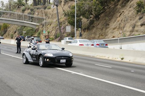 1993 Ford Taurus - A black convertible with police escort drives on a highway, with other vehicles and a pedestrian bridge in the background.