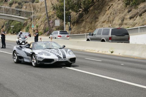 1993 Ford Taurus - A silver Ferrari convertible with a custom decal design drives on a highway, accompanied by police.
