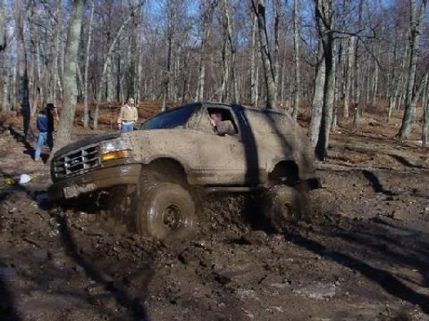 1983 Ford Bronco - This SUV is deep in the mud, with trees and a few people in the background.
